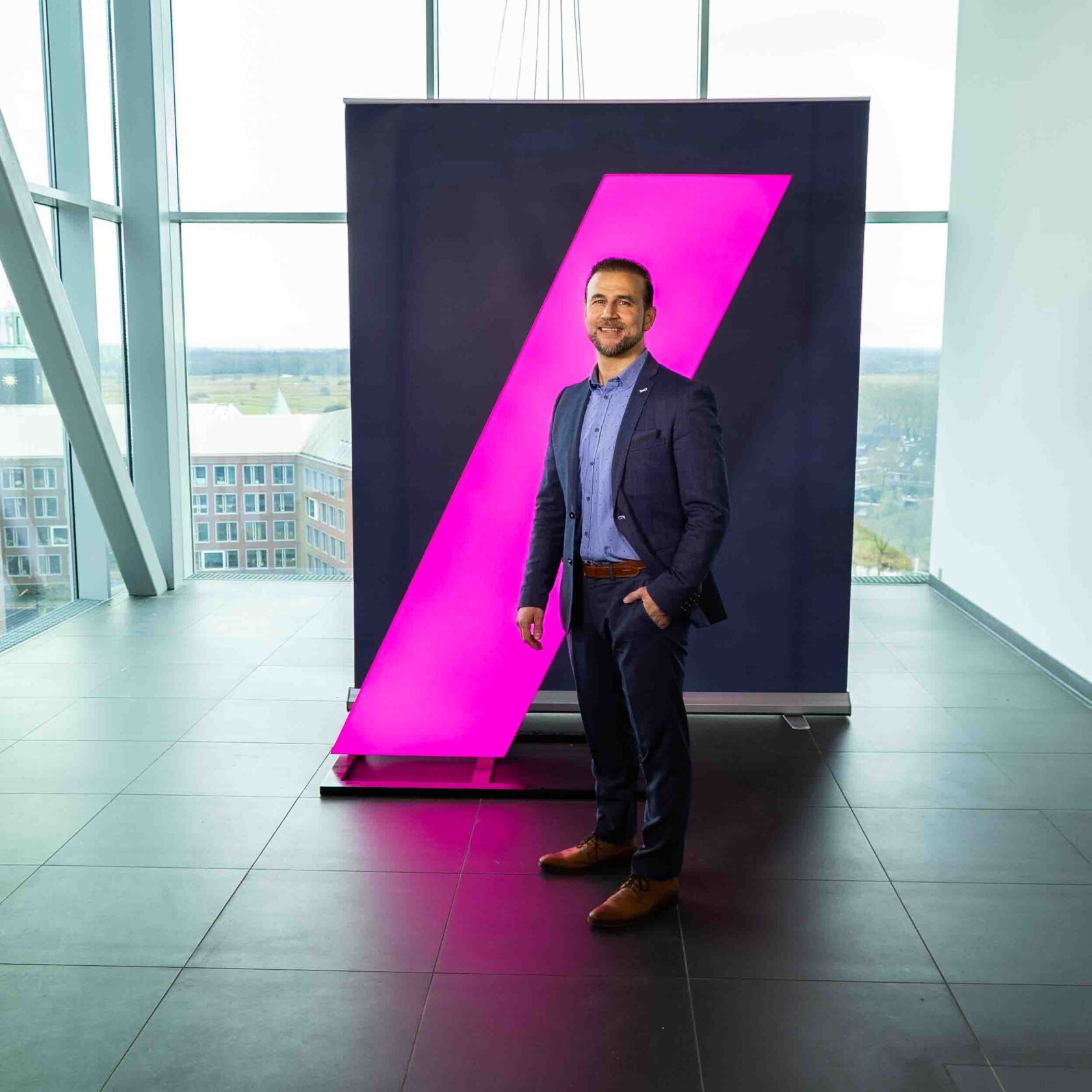 Mustafa Gurel, gekleed in een donkerblauw pak, poseert met een glimlach voor een moderne, zakelijke achtergrond met een opvallende roze lichtinstallatie. Het grote raam biedt uitzicht op een stedelijk landschap, wat zorgt voor een professionele en dynamische sfeer.