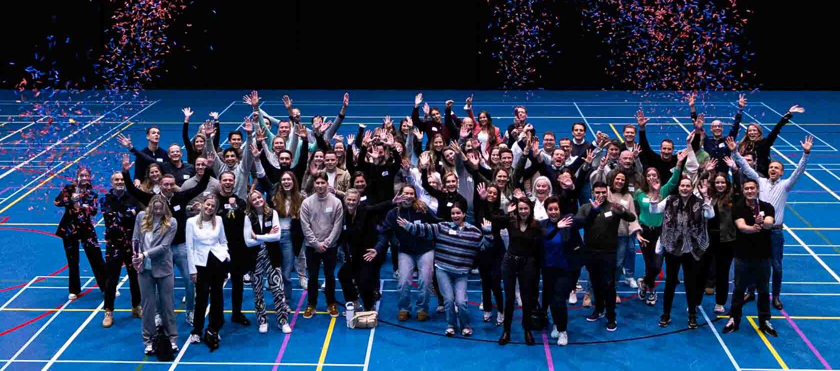 Groepsfoto van enthousiaste medewerkers van Welten Groep in een sporthal, met confetti in de lucht en een groot Welten Groep-logo op de achtergrond.